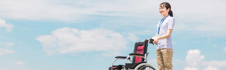介護職員が車椅子を操作する屋外での一場面。晴れた空が背景。